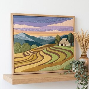 Op de afbeelding: Een ingelijst wandkunstwerk van stof met een landschap van terrasvormige rijstvelden, glooiende bergen en een kleine hut onder een paarse en oranje lucht. Het kunstwerk gebruikt diverse textielsoorten met patronen in groen-, geel- en blauwtinten.