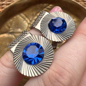Può includere: Un paio di orecchini a clip in metallo color argento con un design circolare a linee radianti e una grande pietra preziosa rotonda, sfaccettata e di colore blu intenso al centro di ogni pezzo.