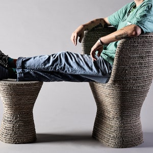K&ouml;nnte beinhalten: Eine Person sitzt auf einem braunen, geflochtenen Stuhl mit einem passenden Fu&szlig;hocker. Stuhl und Fu&szlig;hocker sind aus recyceltem Karton gefertigt.