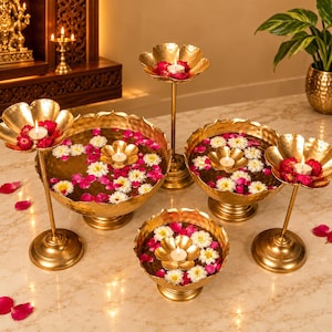 May include: A collection of gold-toned metal bowls and elevated candle holders with scalloped edges. The bowls are filled with water, floating white and pink flowers, and small tea light candles. The metal surfaces have a hammered texture.