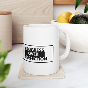 Könnte beinhalten: Ein weißer Keramikbecher mit einer rechteckigen schwarzen Grafik und dem weißen Text "PROGRESS OVER PERFECTION" in der Mitte. Der Becher hat eine glatte Oberfläche, einen geschwungenen Henkel auf der rechten Seite und steht auf einem quadratischen Untersetzer.
