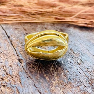 Peut inclure: Une bague dorée présentant un motif de coquillage cauri poli en guise de pièce centrale. La bague possède une finition métallique lisse avec une ouverture horizontale centrale caractéristique d'un coquillage, montée sur un anneau simple.
