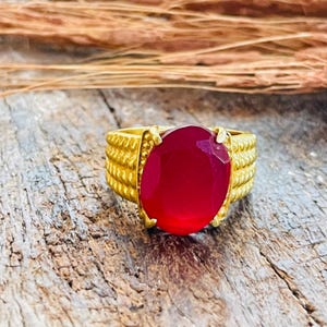 Peut inclure: Une bague en or ornée d'une grande pierre précieuse rouge foncé de forme ovale au centre. L'anneau de la bague présente un design texturé et strié qui fait le tour du doigt, offrant un contraste marqué avec la pierre rouge lisse et polie.