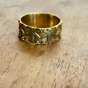 May include: A gold-toned metal band ring featuring a repeating embossed pattern of seven-pointed leaves. The design includes small raised spherical beads along the top and bottom edges of the band, creating a textured, metallic finish.