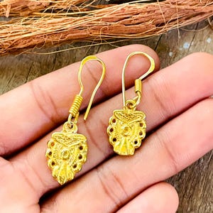 Puede incluir: Un par de pendientes colgantes de tono dorado con un colgante texturizado en forma de escudo que presenta un intrincado patrón en relieve y un detalle circular central. Cada pendiente cuelga de un gancho dorado con un pequeño detalle en espiral.