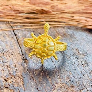 May include: A gold-toned ring featuring a central turtle motif. The turtle shell has a geometric hexagonal pattern, and the ring band displays a textured, dotted surface detail. The metal has a matte finish, and the design is symmetrical.