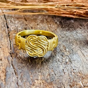 Puede incluir: Un anillo de color dorado que presenta un diseño de nudo central con detalles intrincados y texturizados en la banda. La superficie metálica tiene un acabado pulido, resaltando el patrón trenzado de la pieza central y los elementos decorativos laterales.
