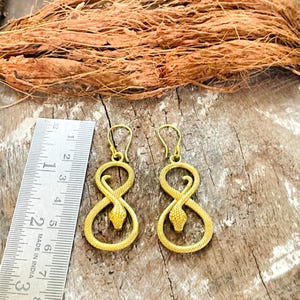 May include: A pair of gold-toned snake-shaped drop earrings featuring a textured scale pattern and a hook closure. A metal ruler is positioned to the left, showing the earrings measure approximately 6.5 cm in total length.