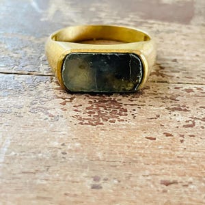 May include: A gold-toned ring featuring a rectangular, dark green, flat-surfaced stone set horizontally in the center. The band is smooth and solid, with a polished finish that contrasts with the textured, opaque appearance of the stone.