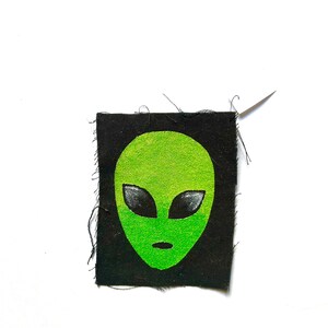 Peut inclure: Un écusson rectangulaire en tissu noir arborant une tête d'extraterrestre vert néon sérigraphiée avec de grands yeux noirs en amande et une petite bouche ovale. L'écusson présente des bords effilochés et une aiguille à coudre argentée fixée.