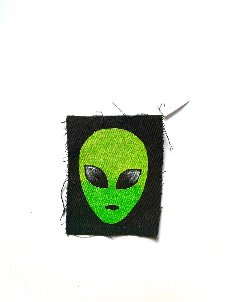 May include: A rectangular black fabric patch featuring a screen-printed neon green alien head with large, almond-shaped black eyes and a small oval mouth. The patch has raw, frayed edges and a single silver sewing needle attached at the top right corner.