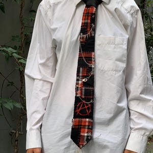 Peut inclure: Une cravate à motif écossais noir et rouge avec des coutures apparentes, ornée de chaînes en métal argenté, de petites épingles à nourrice et d'un symbole anarchiste brodé en rouge. La cravate est portée sur une chemise blanche à manches longues.