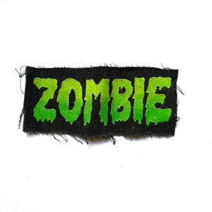 May include: A rectangular fabric patch with a black background and the word ZOMBIE printed in a bright, lime green, dripping font. The edges of the patch are frayed, showing loose black threads along the top, bottom, and sides of the rectangle.