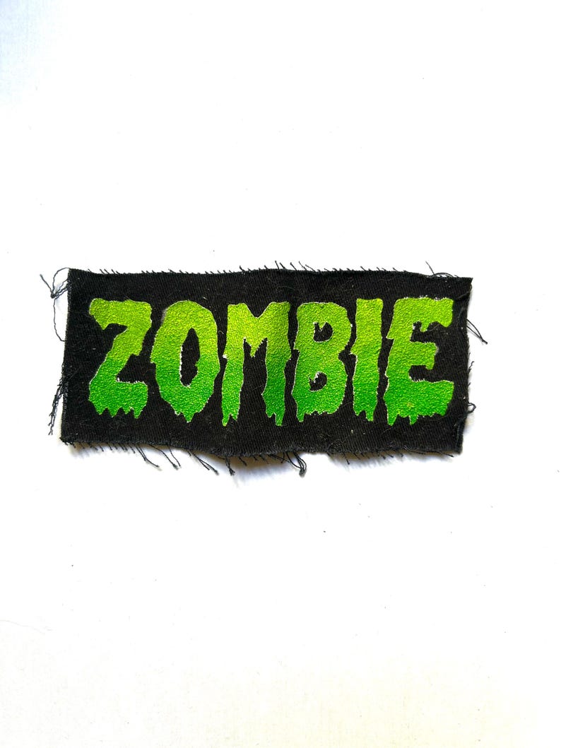May include: A rectangular fabric patch with a black background and the word ZOMBIE printed in a bright, lime green, dripping font. The edges of the patch are frayed, showing loose black threads along the top, bottom, and sides of the rectangle.
