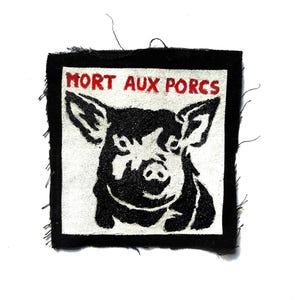 May include: A square fabric patch with a black border and a white center featuring a black graphic illustration of a pig's head. The text "MORT AUX PORCS" is embroidered in red capital letters across the top of the white square area.