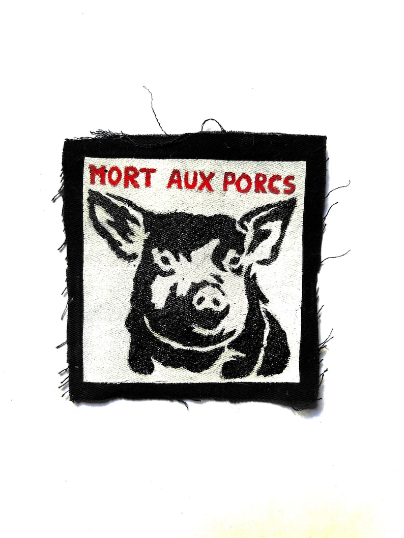 May include: A square fabric patch with a black border and a white center featuring a black graphic illustration of a pig's head. The text "MORT AUX PORCS" is embroidered in red capital letters across the top of the white square area.