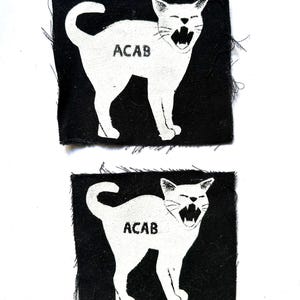 Peut inclure: Deux écussons carrés en tissu noir aux bords effilochés, chacun présentant un motif sérigraphié blanc d'un chat feulant au dos arqué. Le texte ACAB est imprimé en noir sur le flanc de chaque silhouette de chat, créant un contraste marqué.