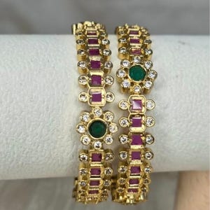 May include: Two gold-tone bangles featuring a repeating pattern of square-cut magenta stones and circular green stones, each surrounded by small clear crystals. The text House of Nilanka is printed in black at the bottom of the image.