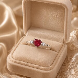May include: A silver-toned ring featuring a central heart-shaped red gemstone flanked by two small, round, clear stones. The ring is displayed inside an open, beige, velvet-lined jewelry box, showcasing the contrast between the metal and the red stone.