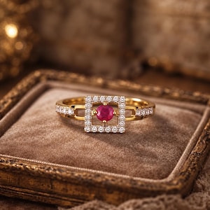 Könnte beinhalten: Ein goldener Ring mit einem zentralen quadratischen Rahmen, der mit kleinen weißen Steinen besetzt ist und einen einzelnen runden roten Edelstein umschließt. Die Ringschiene ist zudem auf beiden Seiten mit einer Reihe kleiner weißer Steine verziert.