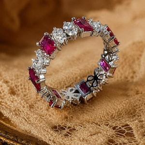 May include: A silver-toned eternity ring featuring an alternating pattern of rectangular-cut deep red stones and pairs of small, round, clear, faceted stones set in a continuous band, showcasing a metallic finish and intricate prong settings.