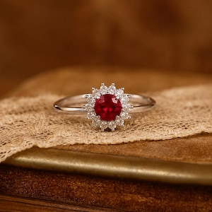 May include: A silver ring featuring a round, deep red gemstone at the center, surrounded by a halo of small, clear, sparkling stones. The band is thin and polished, creating a bright contrast against the vibrant red center stone.