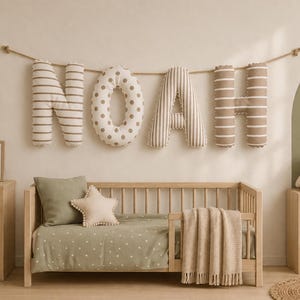 May include: A set of plush, stuffed fabric letters spelling the name NOAH, suspended on a rope. The letters feature alternating brown and white horizontal stripes and a white background with brown polka dots, displayed above a wooden crib.