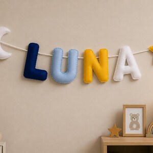 Op de afbeelding: Een decoratieve wandhanger met gevulde stoffen letters die het woord LUNA vormen in marineblauw, lichtblauw, geel en wit. De slinger is voorzien van een witte halve maan aan de linkerkant en een gele ster aan de rechterkant, geregen aan een wit koord.
