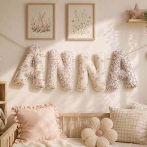 May include: A decorative fabric banner spelling the name ANNA, featuring four plush, stuffed letters in varying floral patterns with white pom-pom trim, suspended on a thin string against a wall above a cot with several throw pillows.