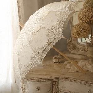 May include: An off-white lace parasol featuring intricate floral embroidery and scalloped edges. The canopy is constructed from delicate fabric panels with detailed needlework, supported by a light-colored wooden handle and central shaft.