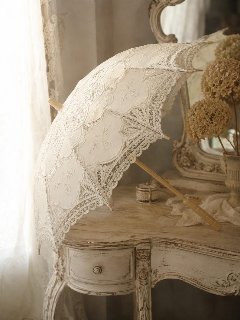 May include: An off-white lace parasol featuring intricate floral embroidery and scalloped edges. The canopy is constructed from delicate fabric panels with detailed needlework, supported by a light-colored wooden handle and central shaft.