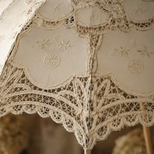 May include: A cream-colored parasol featuring intricate lace panels and scalloped edges. The fabric is adorned with delicate embroidered floral motifs and complex needlework patterns, showcasing a structured frame with a central wooden handle.