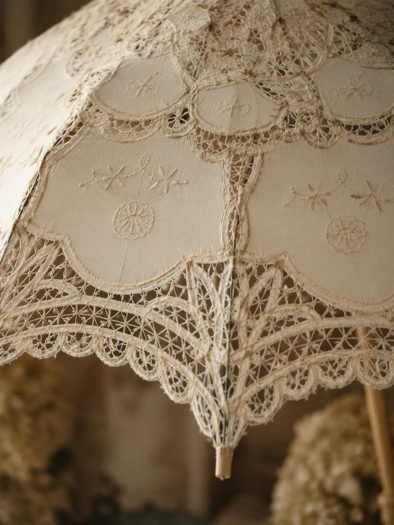 May include: A cream-colored parasol featuring intricate lace panels and scalloped edges. The fabric is adorned with delicate embroidered floral motifs and complex needlework patterns, showcasing a structured frame with a central wooden handle.