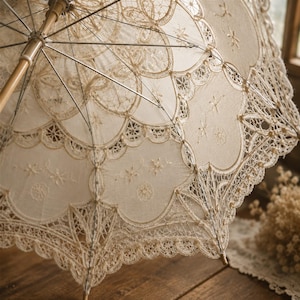 May include: A cream-colored lace parasol featuring intricate floral embroidery and scalloped edges. The canopy is supported by a light-colored wooden handle and thin metal ribs, showcasing detailed needlework patterns throughout the fabric.