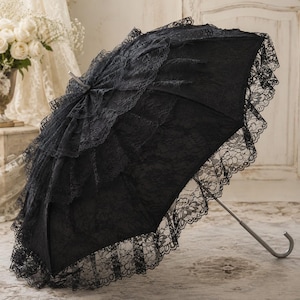 May include: A black umbrella featuring multiple layers of intricate lace ruffles covering the canopy. The umbrella has a curved handle and a central metal shaft, with a wide lace trim along the outer edge of the canopy, creating a layered appearance.