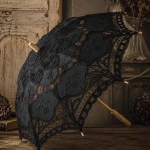 May include: A black lace parasol featuring an intricate floral pattern and scalloped edges. The canopy is supported by a light-colored wooden frame, including a central shaft and a handle with a ribbed grip, finished with a small wooden finial.