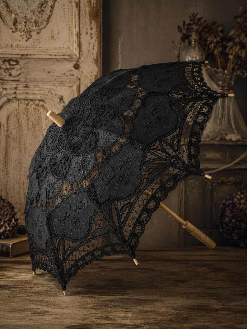 May include: A black lace parasol featuring an intricate floral pattern and scalloped edges. The canopy is supported by a light-colored wooden frame, including a central shaft and a handle with a ribbed grip, finished with a small wooden finial.