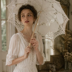 May include: A white lace parasol with intricate floral patterns and scalloped edges, featuring a light-colored wooden handle and central shaft. The parasol is held open, displaying a delicate, semi-sheer fabric construction with detailed embroidery.