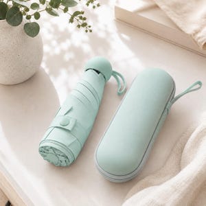 May include: A compact, collapsible umbrella in a solid light mint green colour, featuring a rounded handle with a small wrist strap. Beside it lies a matching, oval-shaped, hard-shell zippered storage case with a fabric wrist strap for portability.