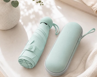 May include: A compact, collapsible umbrella in a solid light mint green color, featuring a rounded handle with a small wrist strap. Beside it lies a matching, oval-shaped, hard-shell zippered storage case with a fabric wrist strap for portability.
