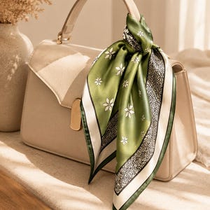 May include: A structured beige handbag with a single top handle, adorned with a square silk scarf tied around the handle. The scarf features a green center with white floral motifs, bordered by a black and white intricate rose pattern trim.