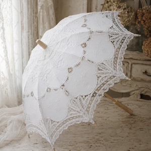 May include: A white lace parasol featuring intricate scalloped edges and detailed embroidery patterns. The canopy is constructed from delicate fabric with openwork lace sections, supported by a light-colored wooden handle and a central wooden finial.