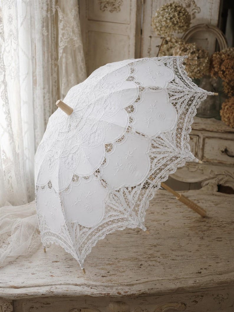 May include: A white lace parasol featuring intricate scalloped edges and detailed embroidery patterns. The canopy is constructed from delicate fabric with openwork lace sections, supported by a light-colored wooden handle and a central wooden finial.