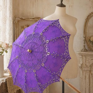 May include: A vibrant purple parasol featuring intricate lace detailing with a circular floral pattern at the center. The canopy is constructed from fabric with elaborate embroidery and scalloped edges, supported by a light-colored wooden handle.