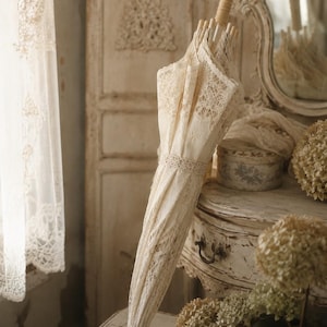 May include: A closed parasol featuring intricate cream-colored lace fabric with detailed floral patterns. The parasol has a light-colored wooden handle and a matching lace band wrapped around the center to keep the canopy securely fastened.
