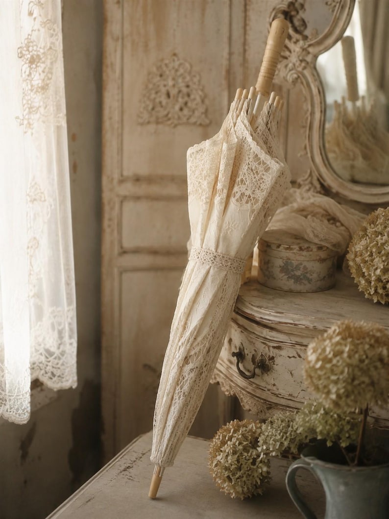 May include: A closed parasol featuring intricate cream-colored lace fabric with detailed floral patterns. The parasol has a light-colored wooden handle and a matching lace band wrapped around the center to keep the canopy securely fastened.