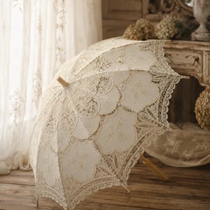 May include: An off-white lace parasol featuring intricate scalloped edges, detailed floral embroidery, and a light-colored wooden handle. The canopy is constructed with layered fabric panels and delicate needlework patterns throughout the design.