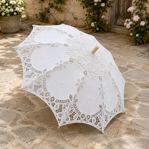 May include: A white lace parasol featuring intricate floral embroidery and scalloped edges. The canopy is constructed from delicate fabric with detailed needlework patterns and a light-colored wooden handle extending from the center top.