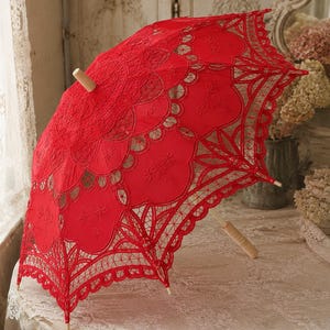 May include: A bright red lace parasol featuring intricate floral embroidery and scalloped edges. The canopy is constructed from detailed needlework patterns, supported by a light-colored wooden handle and a matching wooden tip at the center.
