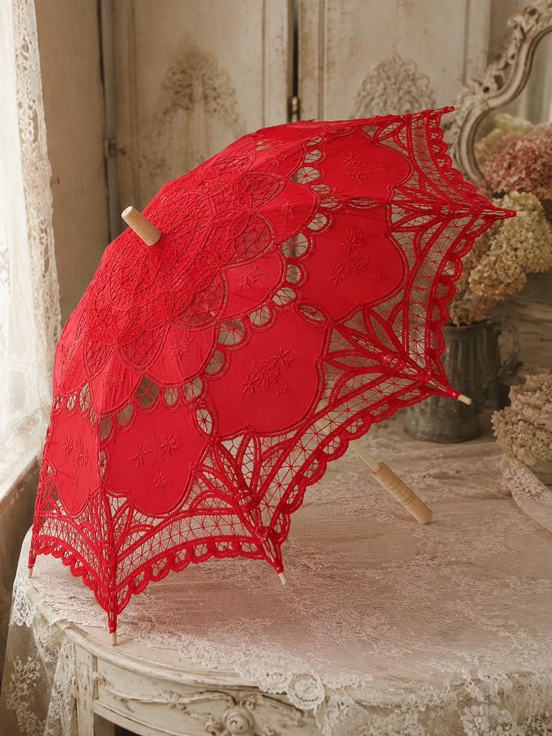 May include: A bright red lace parasol featuring intricate floral embroidery and scalloped edges. The canopy is constructed from detailed needlework patterns, supported by a light-colored wooden handle and a matching wooden tip at the center.